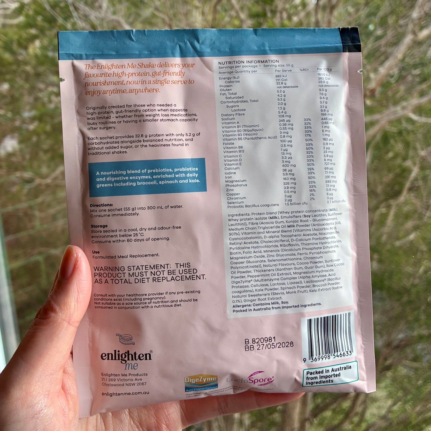 A hand holding the back of the Enlighten Me Chocolate Shake 55g sachet outdoors against a blurred green background, showing the nutrition panel, ingredients, barcode and Enlighten Me logo.
