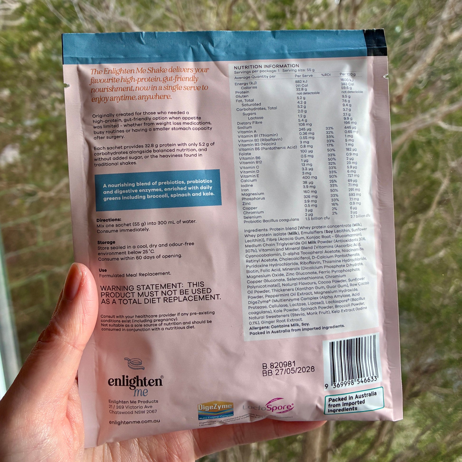 A hand holding the back of the Enlighten Me Chocolate Shake 55g sachet outdoors against a blurred green background, showing the nutrition panel, ingredients, barcode and Enlighten Me logo.