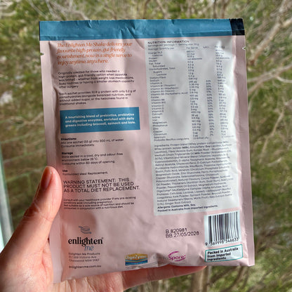 A hand holding the back of the Enlighten Me Chocolate Shake 55g sachet outdoors against a blurred green background, showing the nutrition panel, ingredients, barcode and Enlighten Me logo.