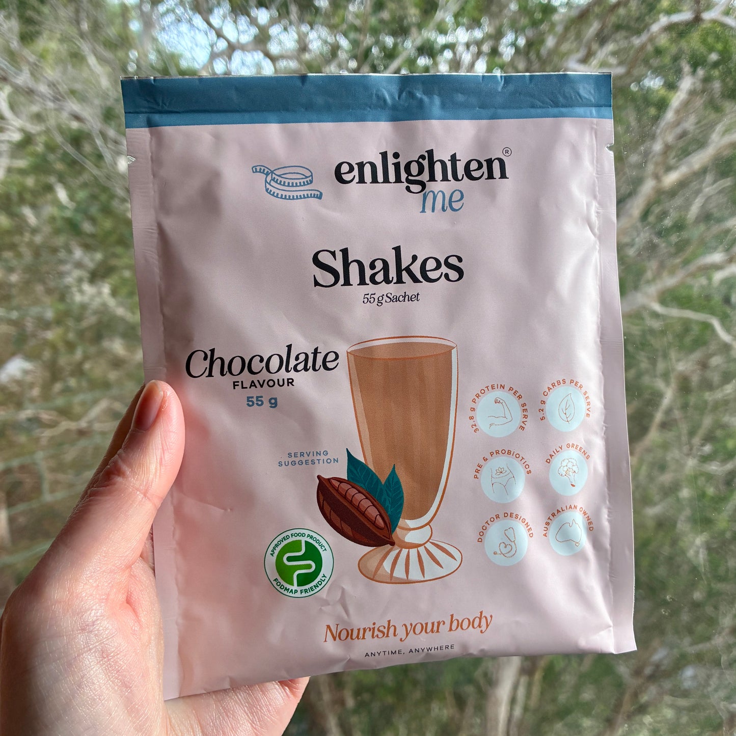 A hand holds a packet of Enlighten Me Shakes Chocolate Flavour 55g sachet in front of a blurred outdoor background of trees.