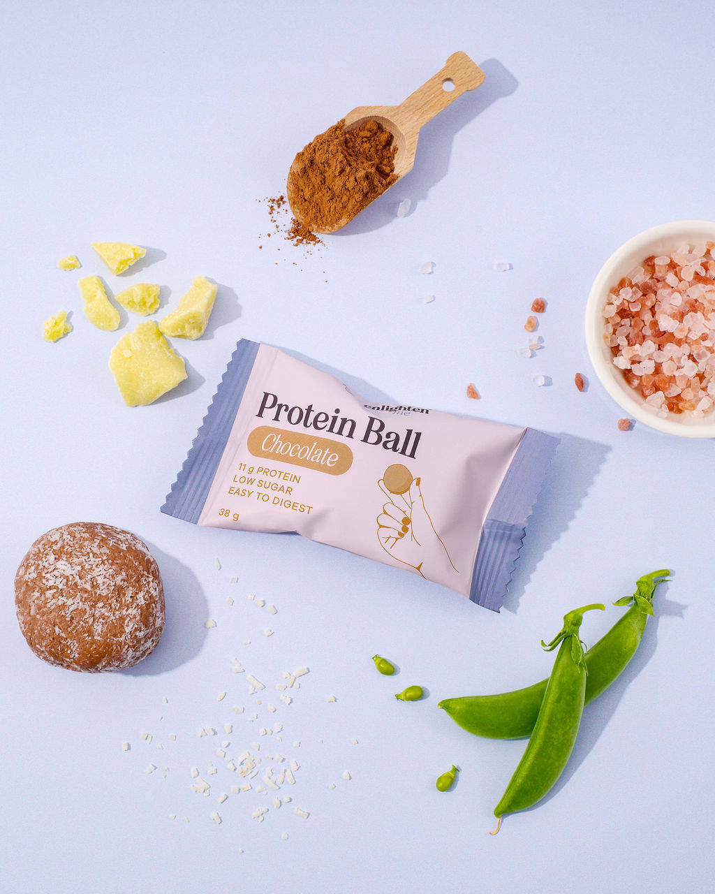 An Enlighten Me Chocolate Protein Ball with its ingredients - cocoa powder, cocoa butter, fine coconut, fresh pea pods and Himalayan salt - beside an unwrapped, coconut-coated protein ball.