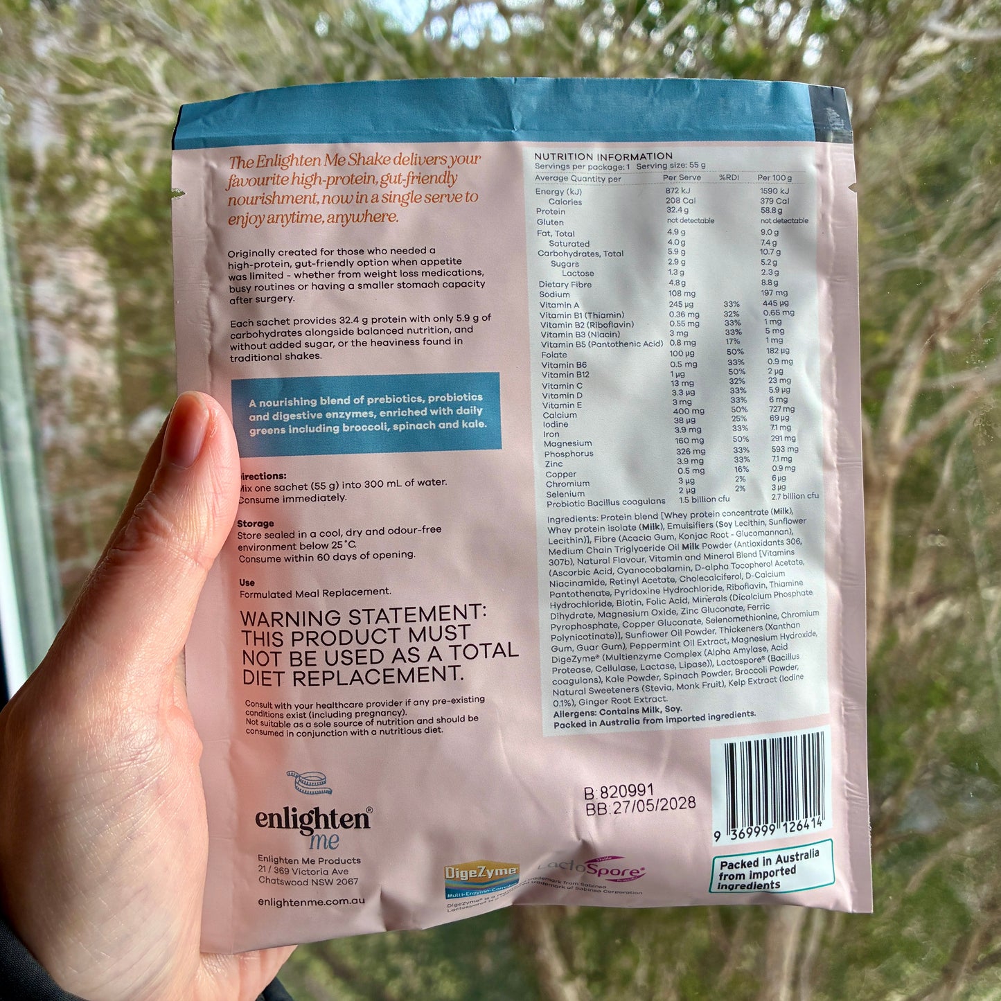 A hand holding the back of the Enlighten Me Vanilla Shake 55g sachet outdoors against a blurred green background, showing the nutrition panel, ingredients, barcode and Enlighten Me logo.