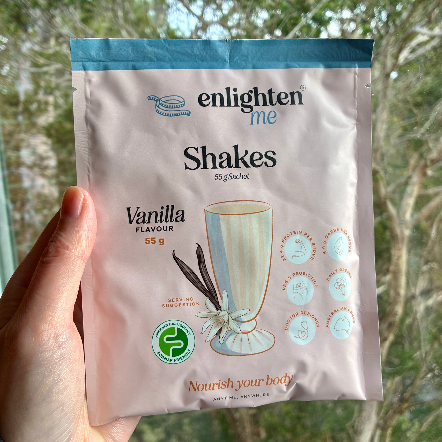 A hand holds an Enlighten Me Vanilla Shake 55g sachet in front of a blurred outdoor background of trees.